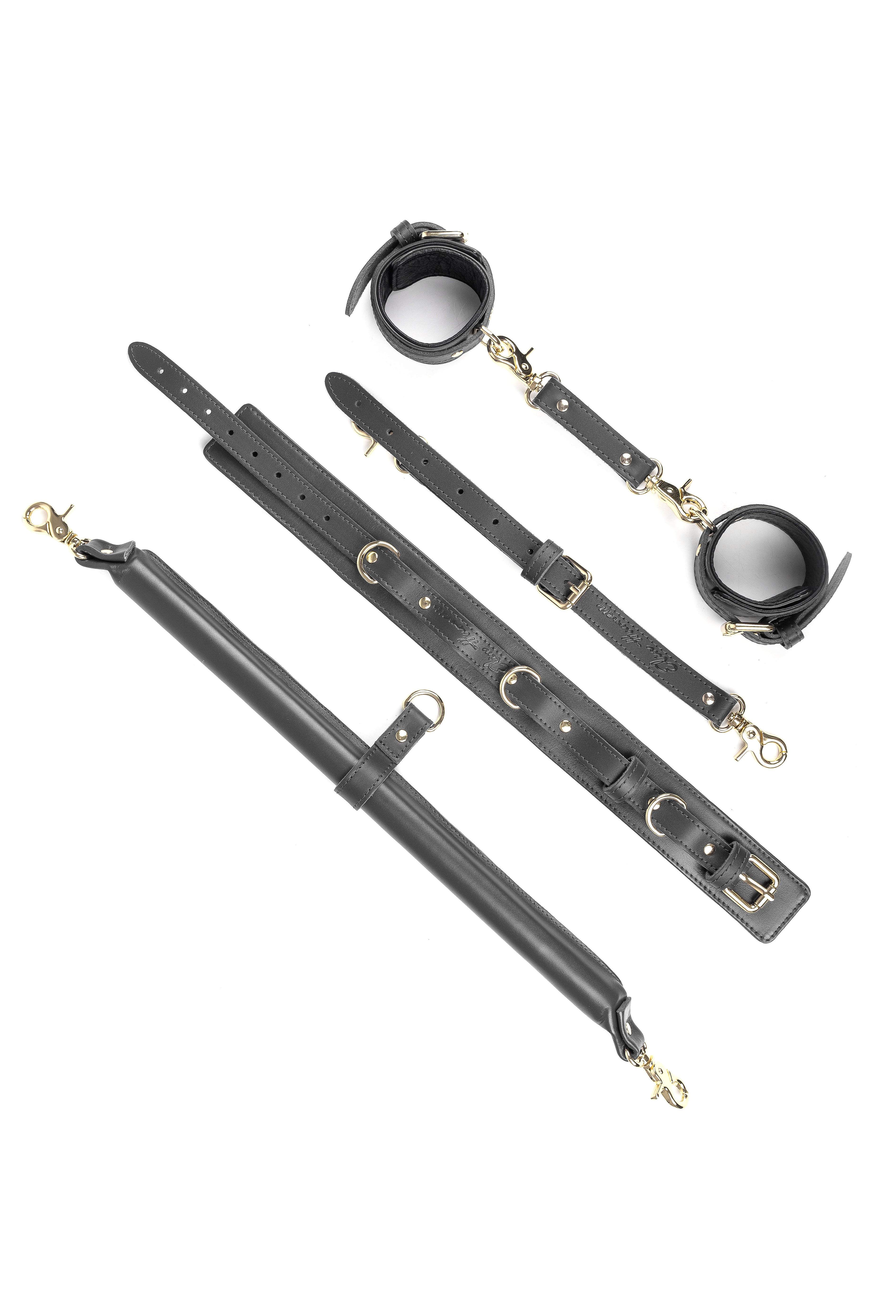 A set of black leather BDSM spreader bar with a wooden base, two carabiner hooks, a D-ring, wrist/ankle cuffs, and a leather collar with a leash.